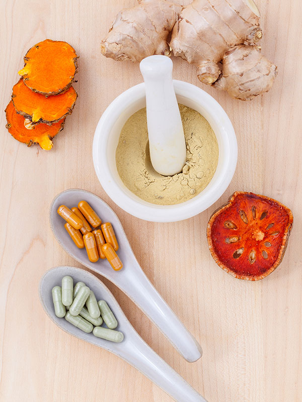 Vitamins, Minerals and Supplements, Ginger, turmeric, dried slime apple, spoons with orange and green capsules, mortar and pestle filled with yellow-beige powder.