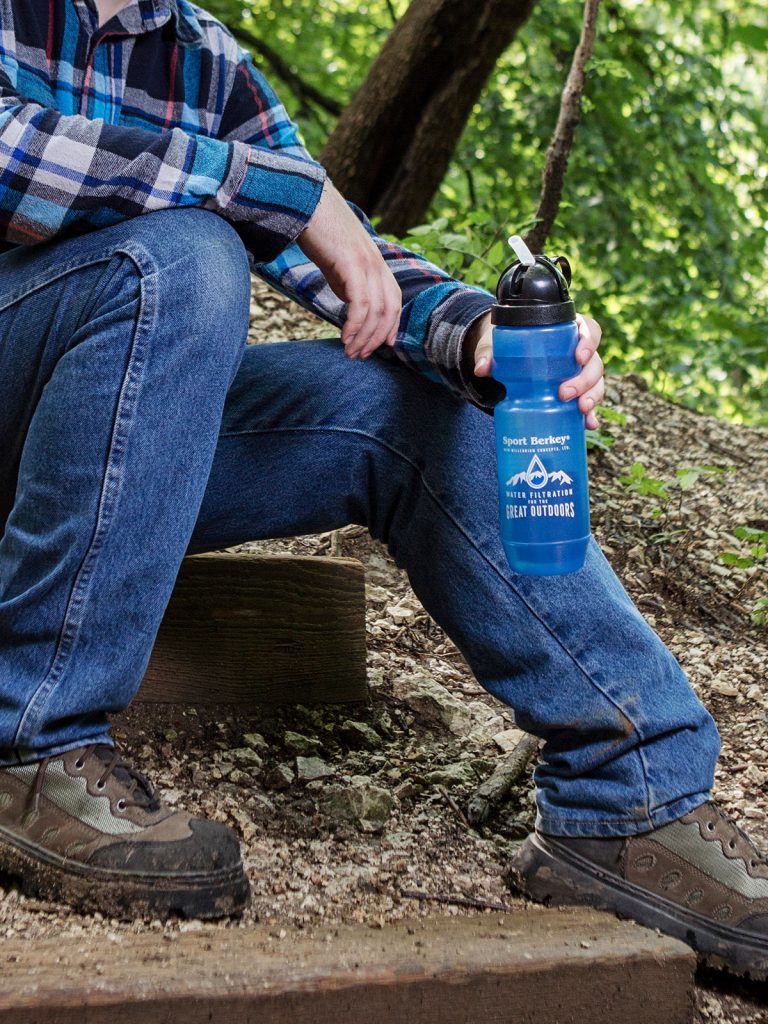 Hombre sentado en un tronco, sostiene Berkey Sport botella de agua azul, sendero